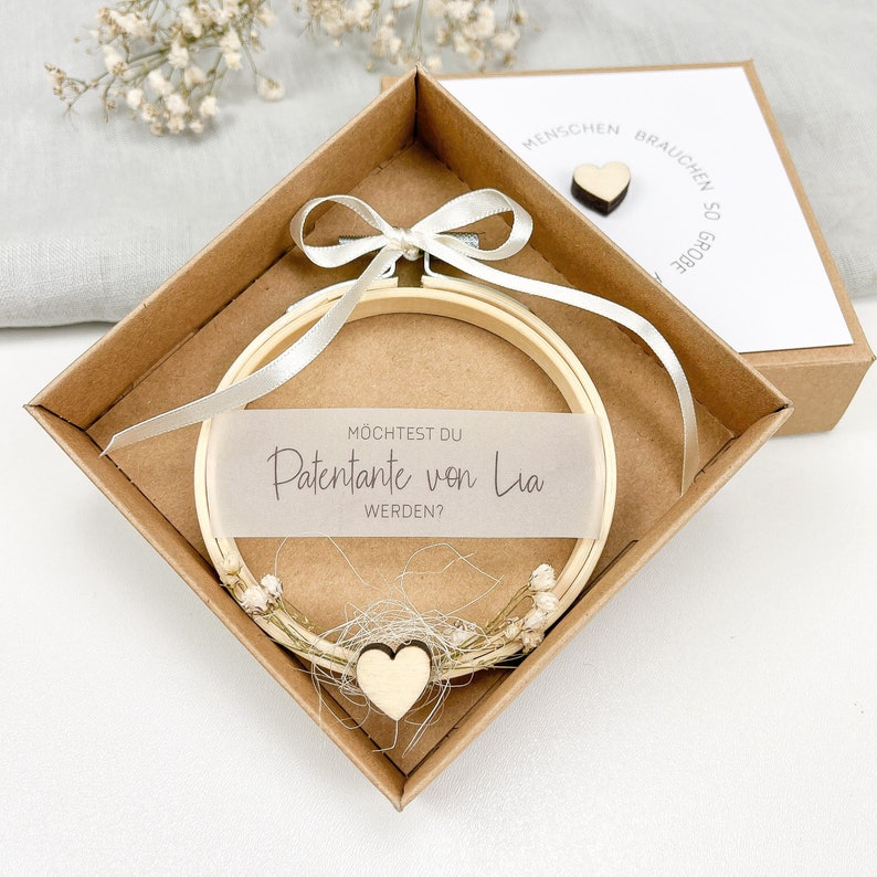 May include: A wooden box with a white ribbon and a wooden embroidery hoop with the text "M&ouml;chtest du Patenante von Lia werden?" inside. The box is open and the hoop is visible.