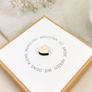 May include: A white card with the text "Kleine Menschen brauchen so gro&szlig;e Herzen wie Deins" in black lettering. A small wooden heart is placed in the center of the card.