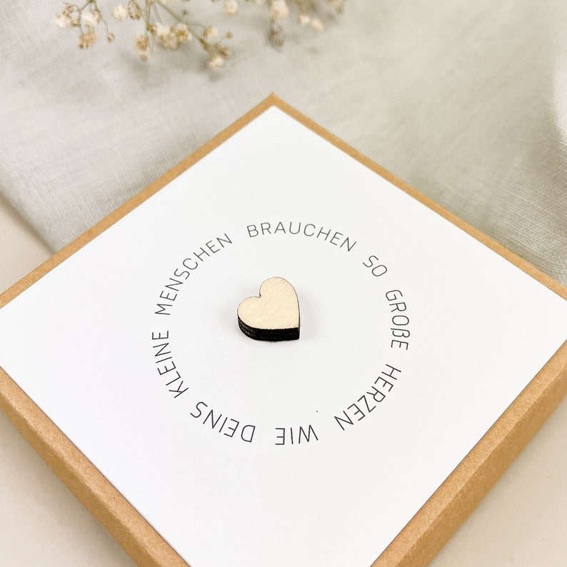 May include: A white card with the text "Kleine Menschen brauchen so gro&szlig;e Herzen wie Deins" in black lettering. A small wooden heart is placed in the center of the card.