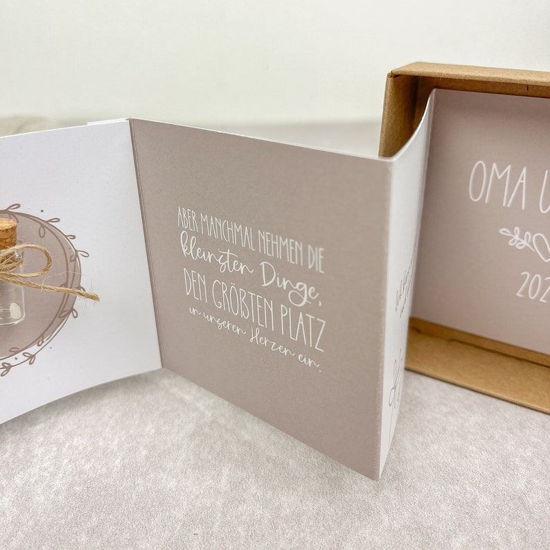 May include: A small, white card with a handwritten message in German. The message reads: "ABER MANCHMAL NEHMEN DIE kleinsten Dinge, DEN GROBTEN PLATZ in unseren Herzen ein." The card is open and a small glass vial with a cork top is visible on the left side of the card. The vial is tied with twine and has a small white object inside.