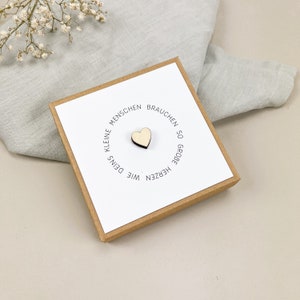 May include: A small wooden heart with the text "Kleine Menschen brauchen so gro&szlig;e Herzen wie Deins" in a circle around it. The heart is on a white card in a brown box.