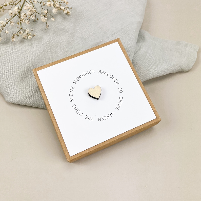 May include: A small wooden heart with the text "Kleine Menschen brauchen so gro&szlig;e Herzen wie Deins" in a circle around it. The heart is on a white card in a brown box.