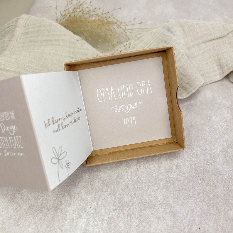 May include: A small, brown box with a white card inside. The card has the text "OMA UND OPA" and the year "2024" printed on it. The card is decorated with a simple floral design and two hearts.
