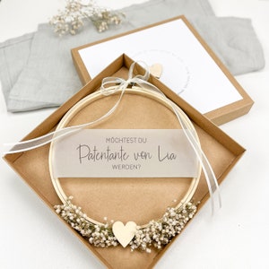 May include: A wooden embroidery hoop with a white ribbon and a small wooden heart. The hoop is in a brown box with a white card that says "M&ouml;chtest du Patenante von Lia werden?" which translates to "Would you like to be Lia's godmother?"