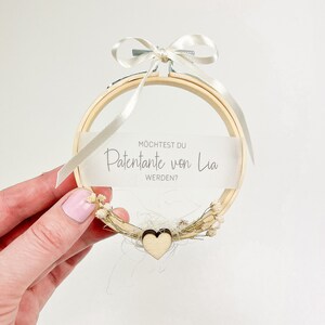 May include: A wooden embroidery hoop with a white ribbon and a small wooden heart. The hoop has a white card with the text "M&ouml;chtest du Patentante von Lia werden?"