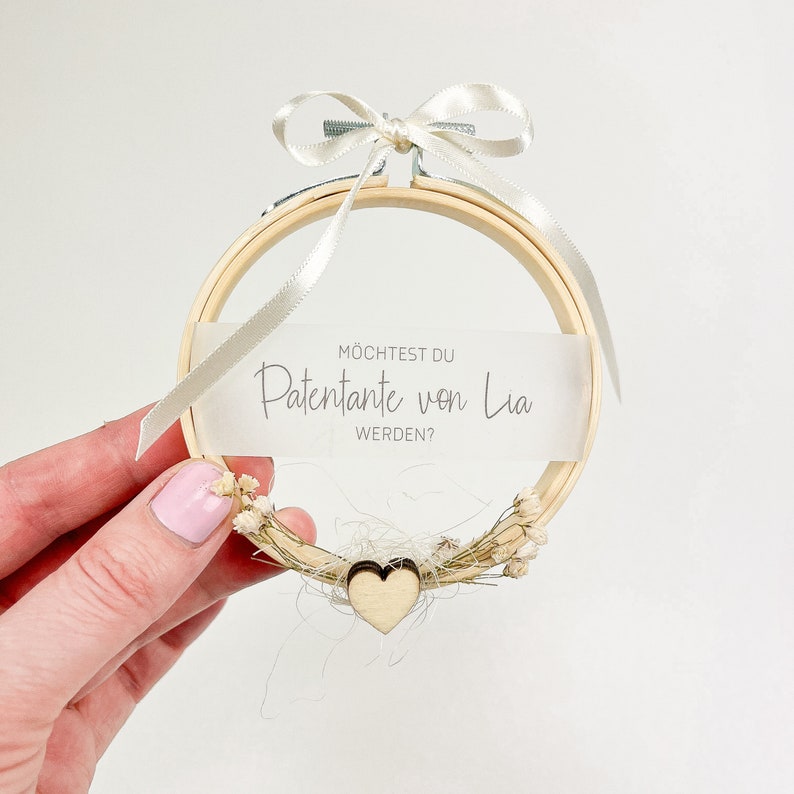 May include: A wooden embroidery hoop with a white ribbon and a small wooden heart. The hoop has a white card with the text "M&ouml;chtest du Patentante von Lia werden?"