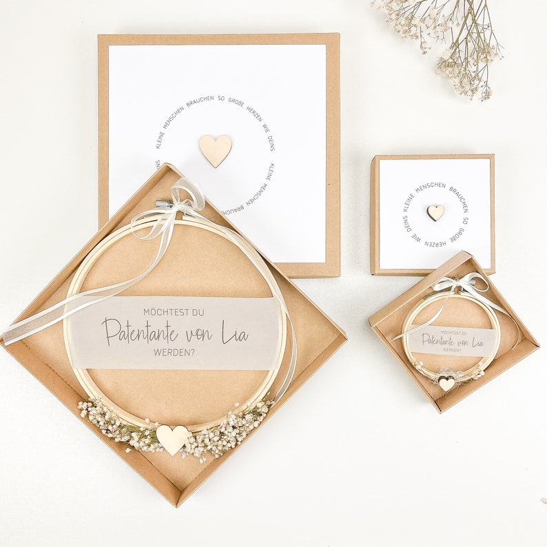 May include: A brown box with a white card inside that says "Kleine Menschen brauchen so gro&szlig;e Herzen wie Deins."  The card has a small wooden heart on it.  The box is open and there is a white embroidery hoop with a white card inside that says "M&ouml;chtest du Patentante von Lia werden?"  The embroidery hoop is decorated with small white flowers.  There is a small wooden heart on the card inside the embroidery hoop.