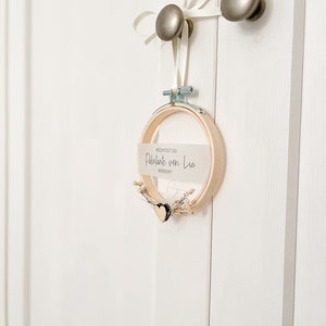 May include: A white card with the text "M&Ouml;CHTEST DU" "Paten/Tante von Lia" "WERDEN?" is attached to a wooden embroidery hoop with a small white heart and dried flowers. The hoop is hanging from a white ribbon tied to a silver door knob.