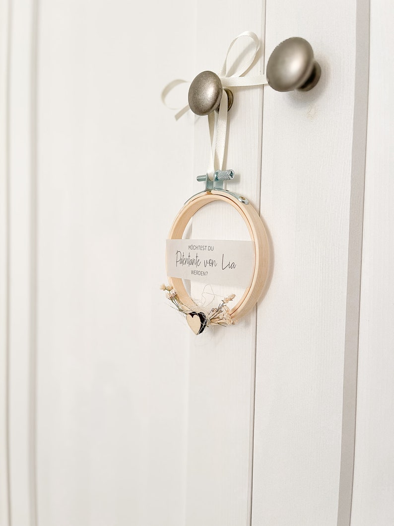 May include: A white card with the text "M&Ouml;CHTEST DU" "Paten/Tante von Lia" "WERDEN?" is attached to a wooden embroidery hoop with a small white heart and dried flowers. The hoop is hanging from a white ribbon tied to a silver door knob.