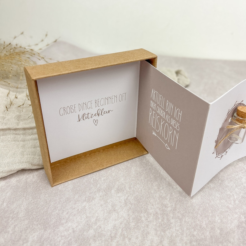 May include: A small, brown box with a white card inside. The card has the text "GROBE DINGE BEGINNEN OFT KLITZKLEIN" with a heart symbol. The box is open and the card is visible. The box is on a white surface.