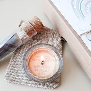 May include: A small glass candle with a white label that reads "Gott Segen Moge Dich Begleiten - Heute, Morgen Und Zu Allen Zeiten". The candle is lit and sitting on a piece of burlap fabric.