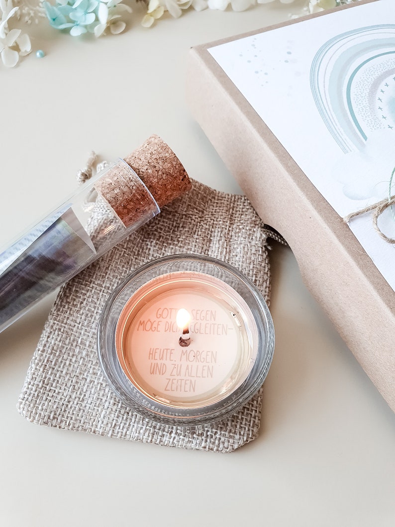 May include: A small glass candle with a white label that reads "Gott Segen Moge Dich Begleiten - Heute, Morgen Und Zu Allen Zeiten". The candle is lit and sitting on a piece of burlap fabric.