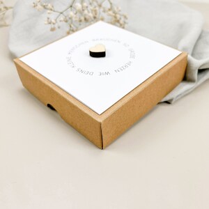 May include: A small brown box with a white card inside. The card has a small wooden heart on it and the text "Wie Deins Kleine Herzen Geboren Brauchen Wir".