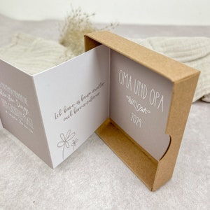 May include: A small, brown cardboard box with a gray interior. The box is open and the inside shows a card with the text "OMA UND OPA" and a small heart. The year 2024 is printed below the text. The card on the left side of the box has the text "Ich kann es kaum erwarten euch kennenzulernen".