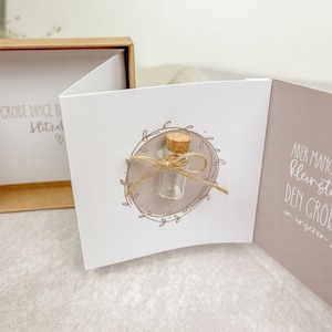 May include: A white card with the text "GROBE DINGE BEGINNEN IMMER MIT KLEINEN SCHRITTEN" and a small glass bottle with a cork stopper tied with twine. The bottle is inside a circle with a floral design. The card is open to reveal a second page with the text "ABER MANCHMAL SIND DIE KLEINSTEN DINGE DIE GR&Ouml;SSTEN IN UNSEREN HERZEN".