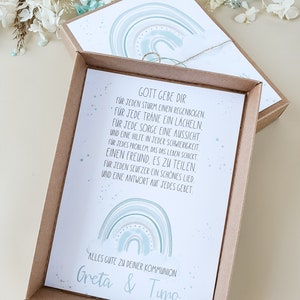 May include: A light brown box with a white card inside. The card features a watercolor rainbow design with the text "Gott gebe dir" and a message about finding strength and support in life. The card also includes the text "Alles gute zu deiner Kommunion Greta & Timo".
