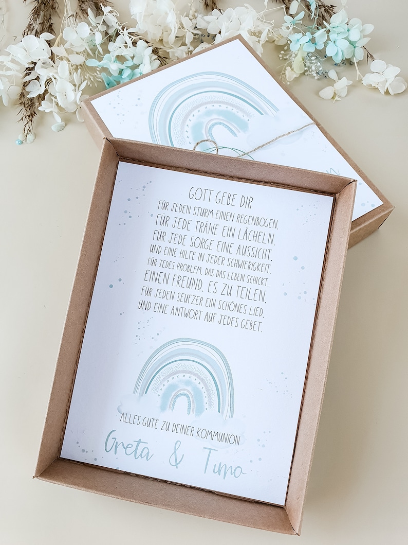 May include: A light brown box with a white card inside. The card features a watercolor rainbow design with the text "Gott gebe dir" and a message about finding strength and support in life. The card also includes the text "Alles gute zu deiner Kommunion Greta & Timo".