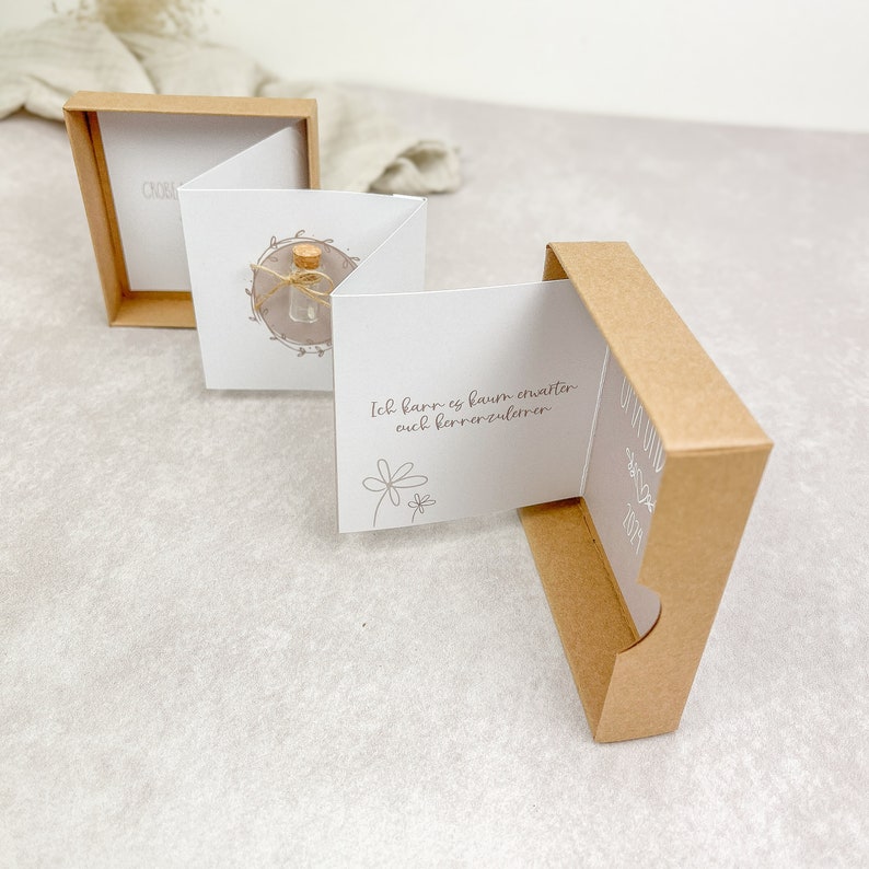 May include: A small, brown box with a white card inside. The card has a small glass bottle with a cork stopper and a handwritten message that reads "Ich kann es kaum erwarten euch kennenzulernen".
