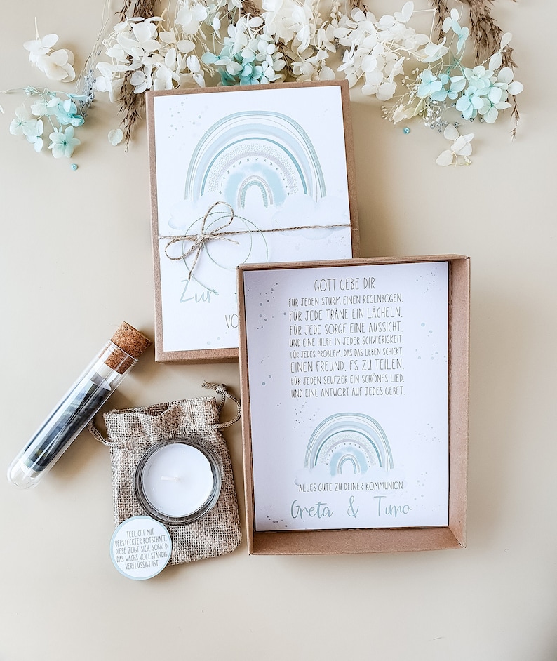 May include: A gift box with a card that says "Gott gebe dir" and a rainbow design. The card also says "Alles gute zu deiner Kommunion Greta & Timo". The box contains a small candle, a small burlap bag, and a test tube filled with small blue and white stones.