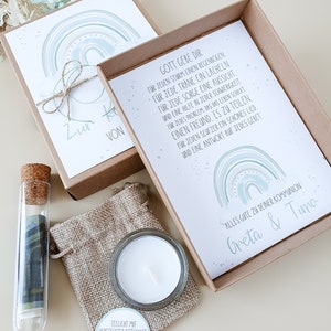 May include: A gift box with a tealight, a small glass vial with a rolled up piece of paper inside, and two cards with a rainbow design and a message about God's blessings. The cards have the text "Zur Kommunion von" and "Gott gebe dir f&uuml;r jeden Sturm einen Regenbogen..." The box is decorated with a brown ribbon and a small burlap bag.