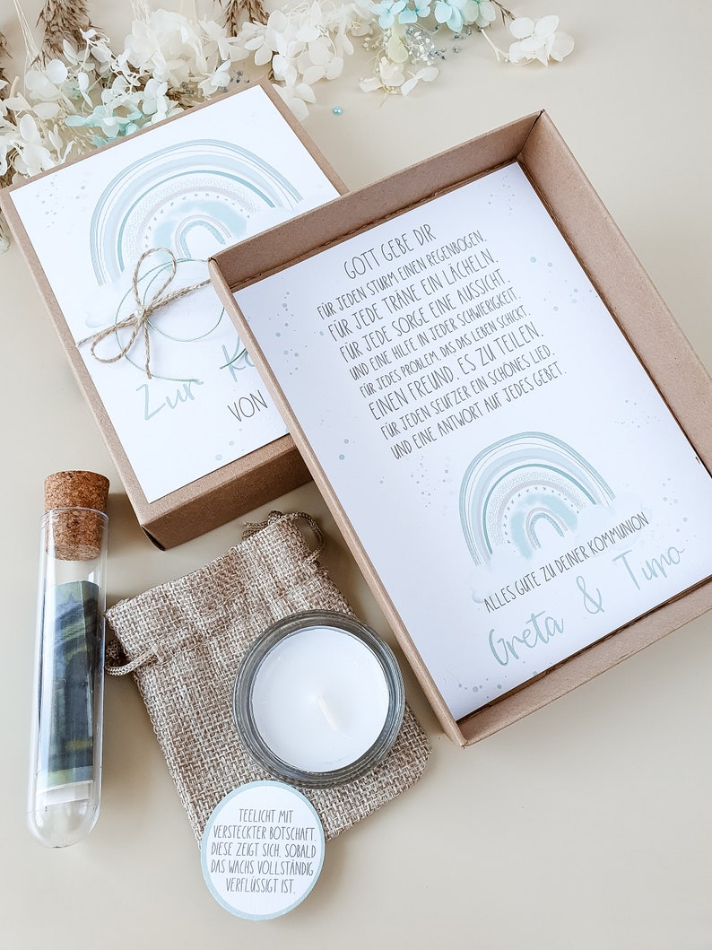 May include: A gift box with a tealight, a small glass vial with a rolled up piece of paper inside, and two cards with a rainbow design and a message about God's blessings. The cards have the text "Zur Kommunion von" and "Gott gebe dir f&uuml;r jeden Sturm einen Regenbogen..." The box is decorated with a brown ribbon and a small burlap bag.