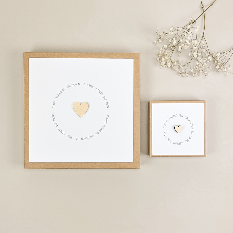 May include: Two brown boxes with white card inserts. The boxes have a circle of text that reads "Kleine Menschen brauchen so gro&szlig;e Herzen wie Deins."  The text is in gray.  A wooden heart is in the center of the circle.