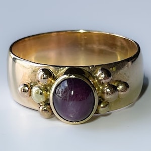 May include: A gold ring featuring a deep purple oval gemstone surrounded by smaller gold beads.