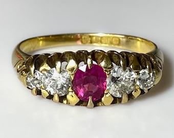 May include: A gold ring featuring a central ruby gemstone flanked by two clear, round-cut diamonds on each side. The ring has a detailed, textured band.