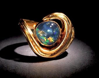 May include: A gold ring featuring a large, oval opal gemstone with a dark blue base and flashes of green and yellow. The ring's band has a swirling, organic design.
