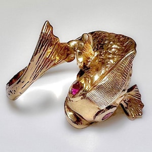 May include: A gold-toned ring featuring a detailed fish design with two pink gemstones for eyes.