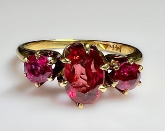 May include: A gold ring with three red gemstones. The center stone is oval shaped and the two side stones are round. The ring is set with claw prongs.
