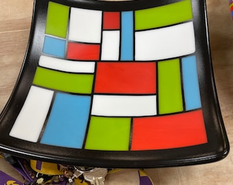 May include: A black ceramic dish with a geometric design in red, green, white, and blue. The dish is slightly curved and has a glossy finish.