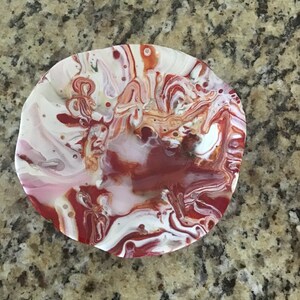 May include: A small, round, white and red resin dish with a swirling, abstract pattern. The dish is made with a resin casting technique and has a glossy finish.
