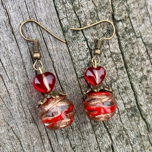 May include: A pair of dangle earrings featuring bronze-toned metal hooks, small translucent red glass hearts, and larger spherical glass beads with swirling red and metallic copper-colored patterns, accented by small bronze-toned metal caps.