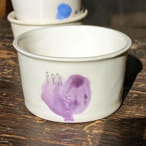 Pode incluir: Um conjunto de taças de cerâmica branca com acabamento liso. A taça da frente apresenta uma mancha estilo aguarela roxa com um pequeno desenho de linhas pretas de três figuras em pé sobre ela. As taças atrás têm superfícies brancas.