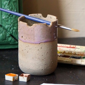 Op de afbeelding: Een keramische penseelhouder met een gespikkelde glazuur en een paarse rand. Een penseel rust in de houder. Er zijn twee kleine aquarelverfpannetjes op een zwart oppervlak.