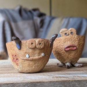 Pode incluir: Duas tigelas pequenas de cerâmica com rostos caprichosos. Uma tigela é marrom com um rosto de porco e a outra é marrom com um rosto de sapo.