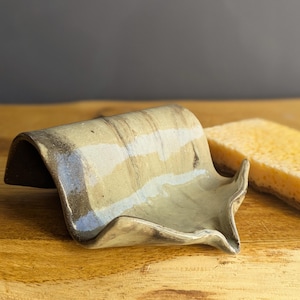 Puede incluir: Una jabonera de cerámica rectangular y curvada con un esmalte brillante de varios tonos que presenta vetas en beige, crema y marrón oscuro. La pieza tiene un borde ligeramente elevado y un pequeño pico, junto a una esponja rectangular amarilla.