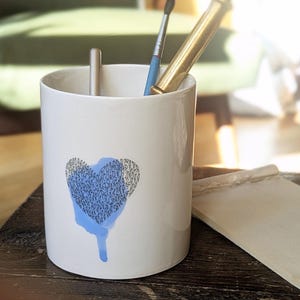 Peut inclure: Pot à crayons en céramique blanche avec un motif de cœur bleu. Le cœur est rempli d'un motif et présente un écoulement bleu. Un petit bloc-notes et des ustensiles d'écriture sont à côté du pot à crayons.