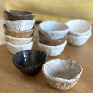 Może przedstawiać: Kolekcja małych, ręcznie robionych ceramicznych misek w różnych kolorach i fakturach. Miski są ułożone i ułożone na drewnianej powierzchni, prezentując ich unikalne kształty i wykończenia. Paleta kolorów obejmuje biel, brąz i czerń.