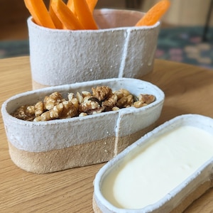 Puede incluir: Un conjunto de tres fuentes de cerámica hechas a mano. Una fuente contiene palitos de zanahoria, otra nueces y la tercera un dip cremoso blanco. Las fuentes tienen un diseño de dos tonos con una sección superior blanca y una base beige.