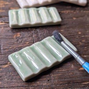 Könnte beinhalten: Eine rechteckige Pinselablage aus Keramik mit glänzend hellgrüner Oberfläche und fünf parallelen Rillen. Die Oberfläche ist glatt und reflektierend. Ein dünner Pinsel mit dunklen Borsten und blauem Stiel liegt auf der gerillten Oberfläche.