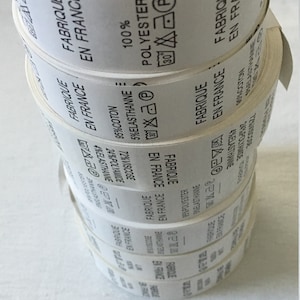 May include: A stack of white fabric labels with black text. The labels read "FABRIQUE EN FRANCE" and include fabric composition percentages and care symbols. The labels are rolled up.