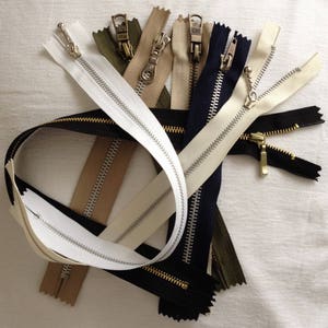 May include: Assortment of different coloured and metal zipper closures. The zippers are arranged in a pile and include white, beige, black, and green.
