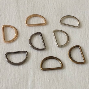 May include: Assortment of eight metal D-rings in various finishes, including gold, silver, and bronze. The rings are all the same size and shape, and they are arranged in a random pattern on a white background.