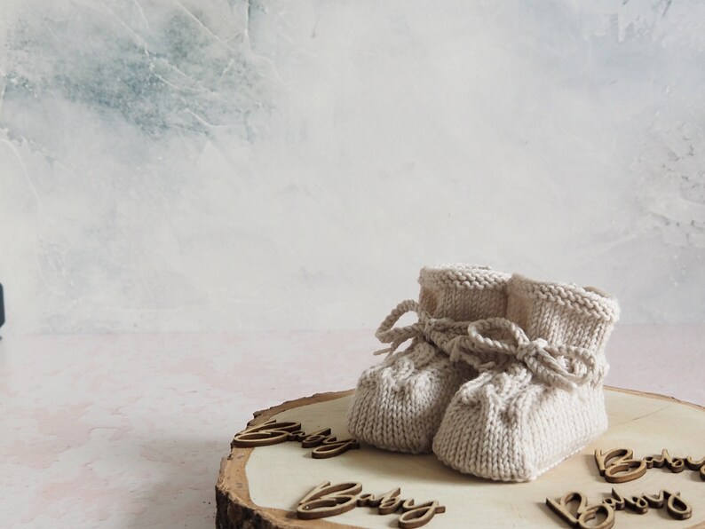 K&ouml;nnte beinhalten: Ein Paar gestrickte Babyschuhe in Cremefarbe mit einer Schleife auf der Vorderseite. Die Schuhe liegen auf einer Holzscheibe mit dem Wort "Baby" darauf.
