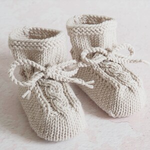 Zart beige Babyschuhe aus Wolle von Hand gestrickt, mit Zopfmuster und Kordeln, die zu Schleifen gebunden sind. Sie wirken sehr natürlich und gemütlich.
