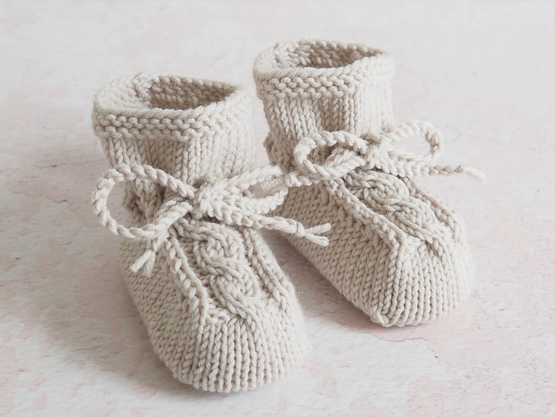 Zart beige Babyschuhe aus Wolle von Hand gestrickt, mit Zopfmuster und Kordeln, die zu Schleifen gebunden sind. Sie wirken sehr natürlich und gemütlich.