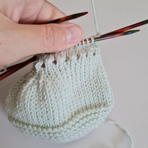 May include: A light green knitted hat being worked on with two knitting needles. The hat is being held in a hand with the yarn attached to the needle.