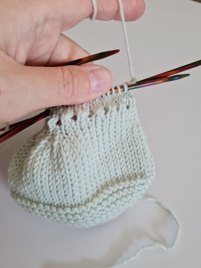 May include: A light green knitted hat being worked on with two knitting needles. The hat is being held in a hand with the yarn attached to the needle.